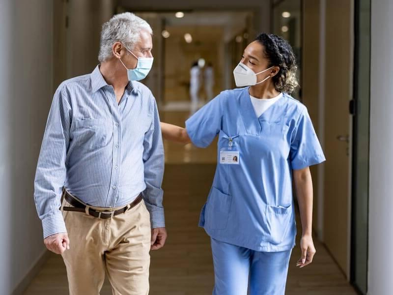 Masked doctor talking with senior patient walking down hallway